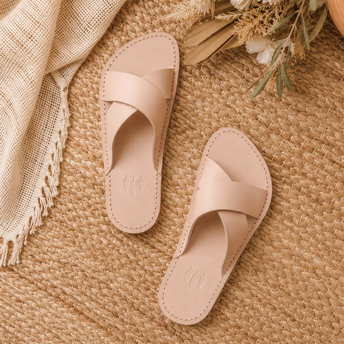 Crema | Cream-colored leather flat sandals with debossed Portland leather tree logo set on a woven mat and bouquet of flowers